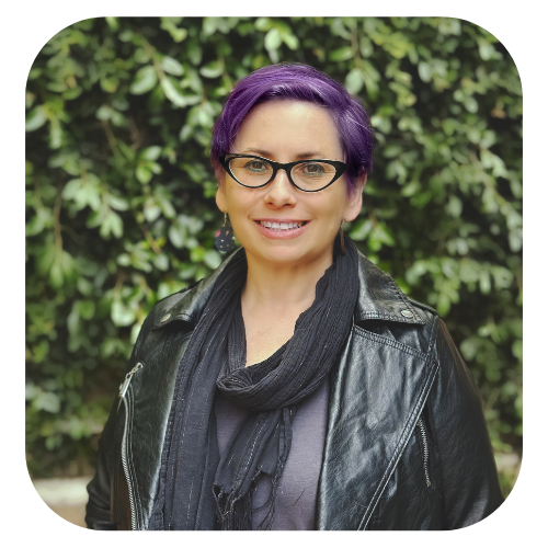 Stacy Jill Calvert is in a black jacket and scarf, with purple hair and black grasses against a wall of green leaves