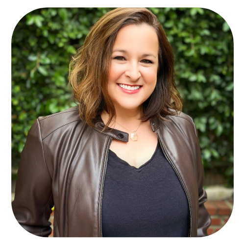 jaclyn Chessen is wearing a brown jacket, has brown hair and is smiling at the camera in front of a wall of green leaves